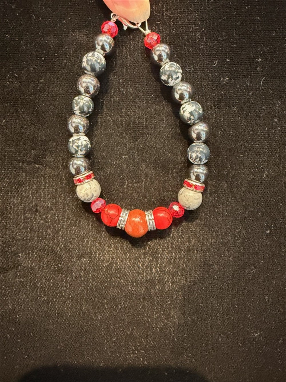 The Hemitite, Snowflake obsidion, picasso jasper, red plastic, red jasper bracelet by Redemption Jewelry features gray, silver, white, and red beads—including a large central red bead—displayed on black fabric.