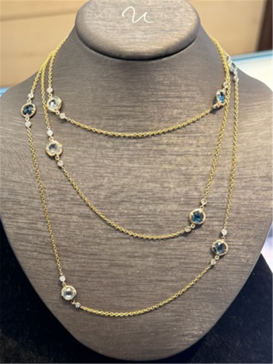 The Ryan Gems Multi Shade Blue Topaz Long Necklace features 14k yellow gold links and sparkling blue topaz stones, elegantly displayed on a gray necklace stand.