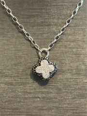 The Ryan Gems Black and White Diamond Clover Pendant in 14k white gold hangs from a silver chain, shown against a textured dark background.