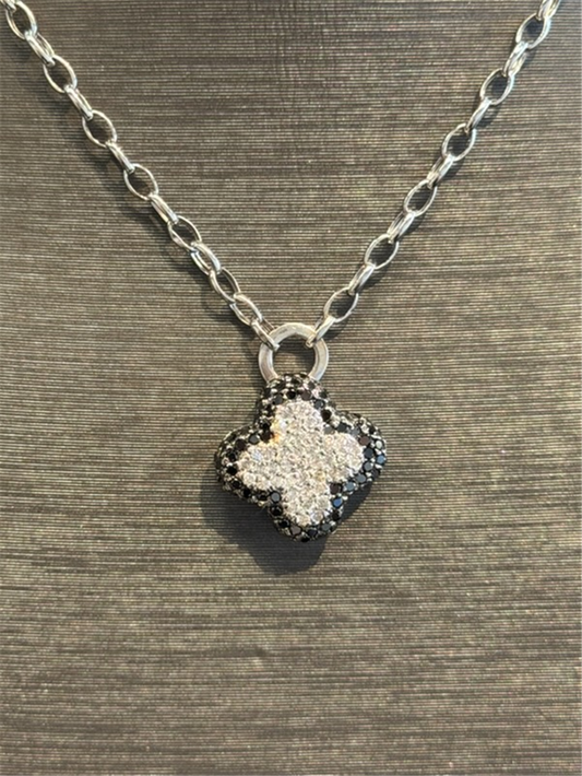 The Ryan Gems Black and White Diamond Clover Pendant in 14k white gold hangs from a silver chain, shown against a textured dark background.