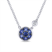 The Gabriel & Co Sterling Silver Blue Sapphire Cluster & Diamond Pendant Necklace, part of the esteemed Lusso Color Collection, features a delicate circular pendant adorned with blue sapphires arranged in a floral pattern, and is complemented by a single small white diamond set on the chain.