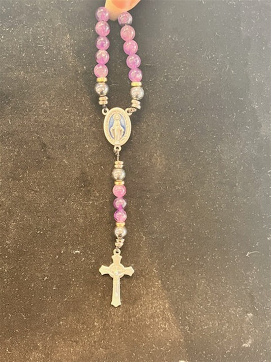 A hand holds the Redemption Jewelry SR001 Small Amethyst & Hematite Rosary, featuring purple beads, a silver Virgin Mary medallion, and a silver crucifix against a dark background.