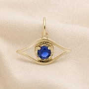 Evil Eye Charm- Gold Plated by Rembrandt Charms, featuring a textured border and central blue gemstone, crafted as an Evil Eye Protection Charm and displayed on a cream-colored fabric background.