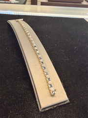 The Ryan Gems Diamond and Princess Cut Sapphire Tennis Bracelet in 14k gold, featuring alternating blue sapphires and diamonds, is displayed on a gray stand against a black velvet background.