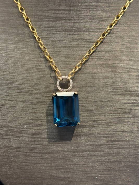 The Ryan Gems London Blue Topaz Statement Pendant, crafted in 14k white and yellow gold, features a rectangular topaz set in a diamond-accented clasp, elegantly displayed on a textured gray surface.