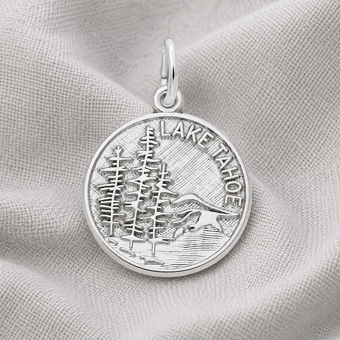 Here’s a revised version using your product data: The Mountain Scene - Lake Tahoe - Sterling Silver Charm by Rembrandt Charms features an engraved round pendant with pine trees, a lake, and mountains, along with the text “LAKE TAHOE.” Set against a soft, light-colored fabric backdrop, this charm is the perfect jewelry piece for nature lovers and an ideal travel keepsake for collectors.