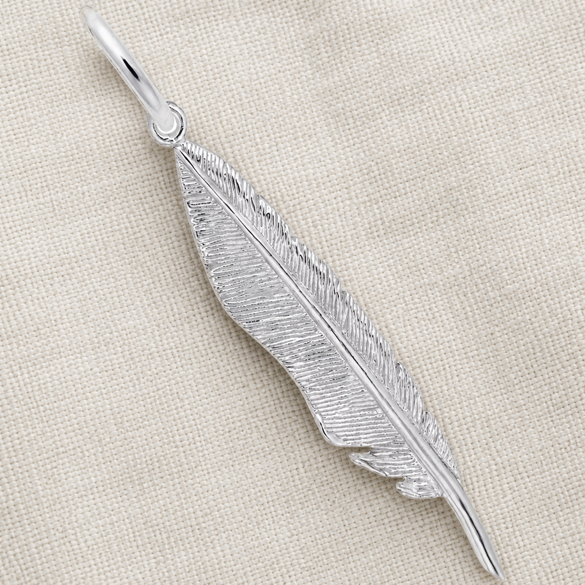 The Feather Charm - Sterling Silver by Rembrandt Charms features a delicately etched, feather-shaped design resting on a light beige, textured fabric background, making it a striking piece of symbolic jewelry.