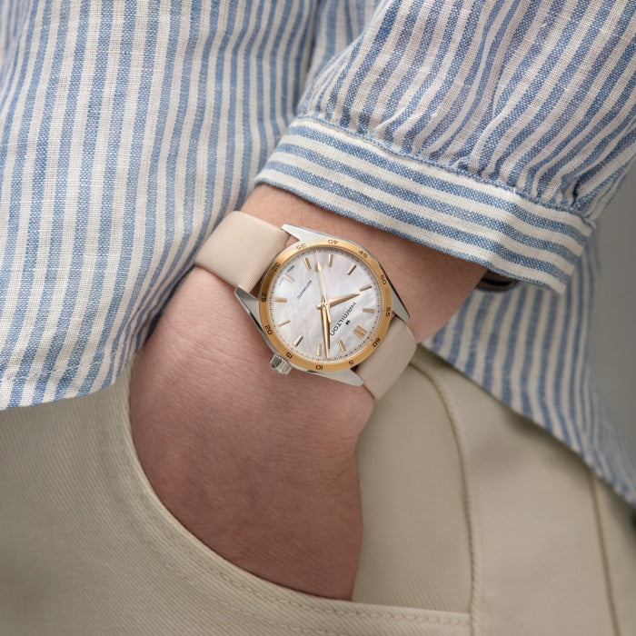 A person wearing a Hamilton Mother of Pearl Automatic with a silver and gold-toned round face, featuring an automatic movement and a white dial, along with a beige leather strap. The person's hand is tucked into the pocket of light-colored pants, and they are wearing a blue and white striped shirt.