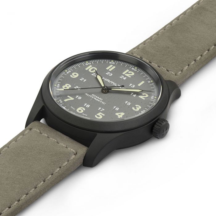 A close-up image of the Hamilton Khaki Field Titanium Automatic 38mm wristwatch, featuring a matte black metal case and a grey suede strap. The watch face is black with white Arabic numerals, military time markings, and luminescent hands and numerals. This 38mm case design exemplifies rugged elegance in military watches.
