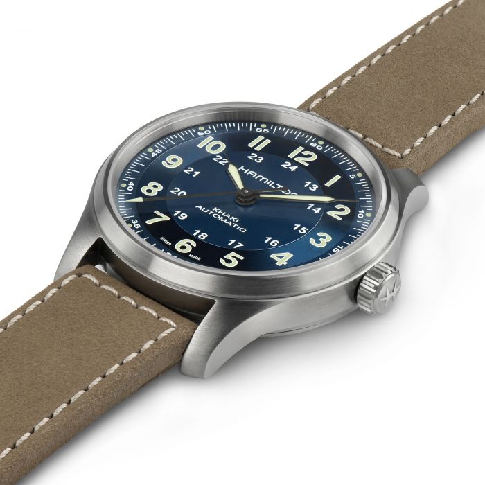 The image showcases a close-up view of the Hamilton Khaki Field Titanium Automatic Watch, which features a silver casing, blue dial, white numerals and markers, and a beige leather strap. This 42mm watch exudes a classic military design with both 12-hour and 24-hour time formats.