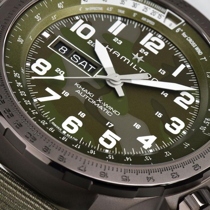 Close-up image of the Hamilton Khaki Aviation X-Wind Automatic Watch. The timepiece features a green camouflage-patterned dial with large white Arabic numerals, a date display at 9 o’clock, and silver hour and minute hands. The detailed markers on the bezel add to its distinct look, complemented by a green strap.