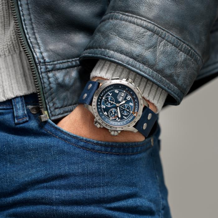 A person wearing a blue leather jacket and jeans has their hand in their pocket, displaying a Hamilton Khaki Aviation X-Wind Automatic Chronograph Watch. The high-performance timepiece, featuring a blue strap and silver casing with multiple sub-dials and side buttons, epitomizes precision.