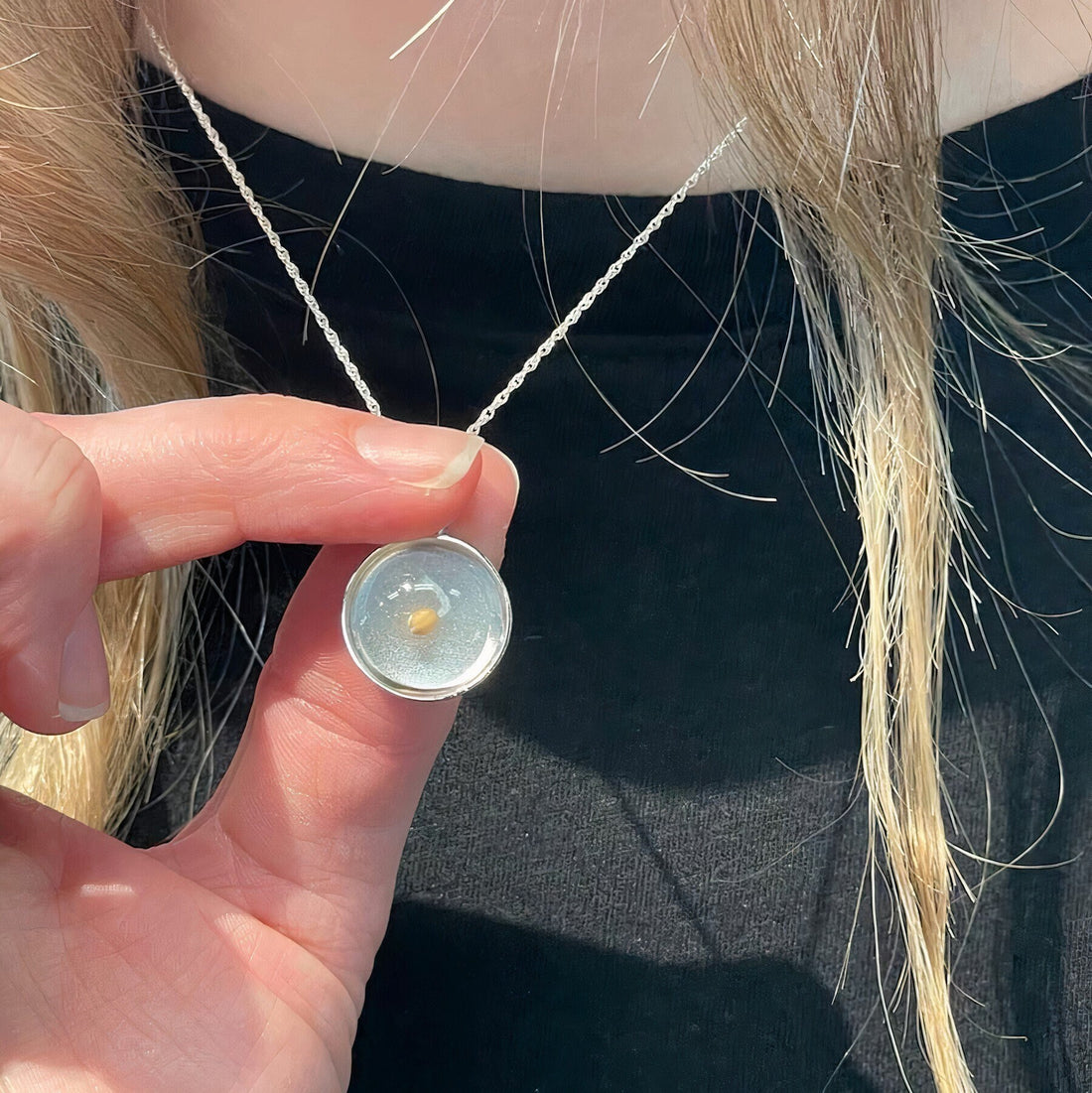 A person with light brown hair is holding the Avonlea Mustard Seed Charm on Rope 20" Chain Necklace in Sterling Silver. The circular pendant features a translucent center that encases a small, light brown object. The person is dressed in a black top.