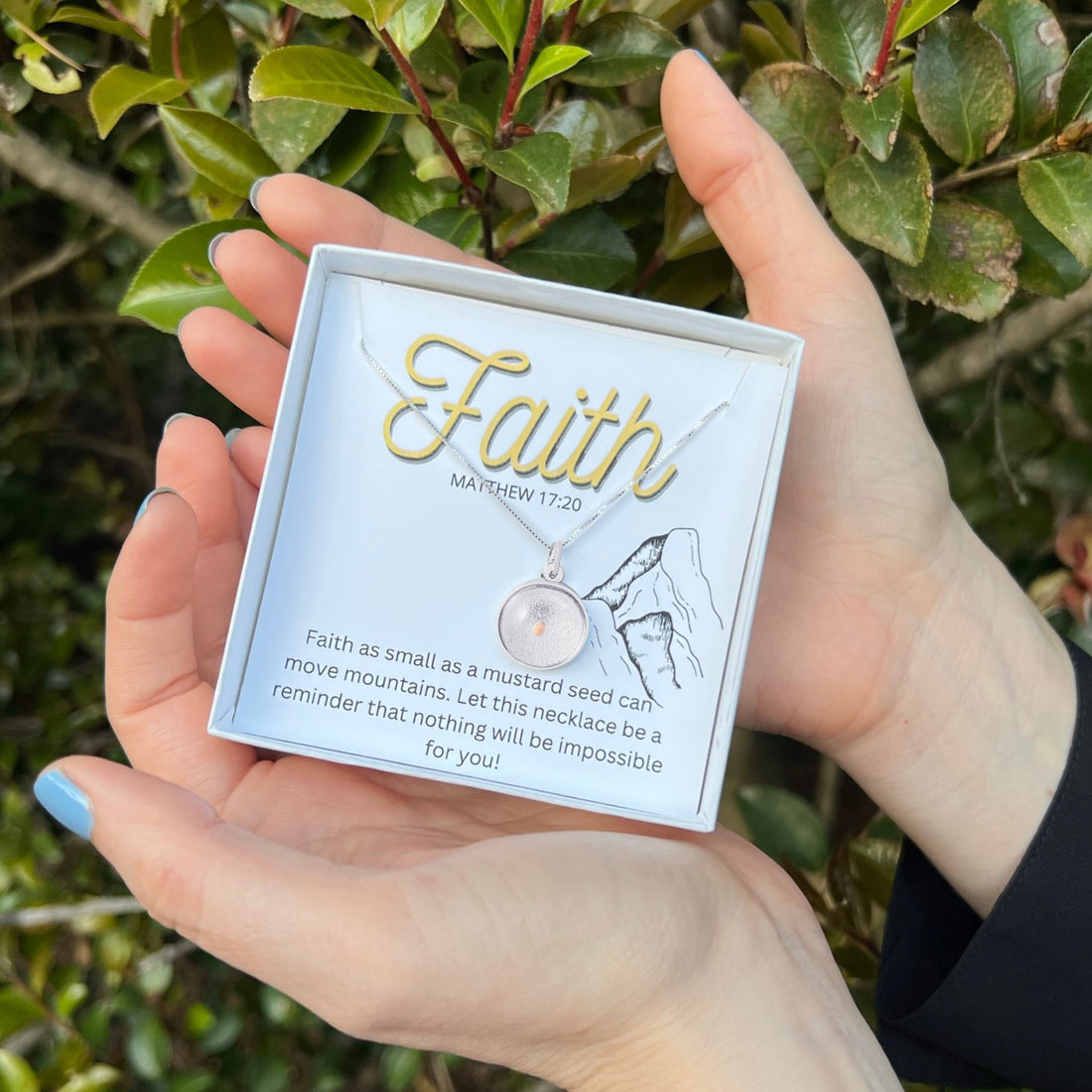 A person holds an Avonlea jewelry box containing a Mustard Seed Charm on Box 18" Chain Necklace Sterling Silver. The box features the word "Faith" in gold lettering and has the Bible verse, Matthew 17:20, about faith moving mountains. Green foliage is in the background, highlighting this beautiful piece of faith jewelry.