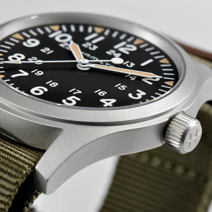 Close-up image of the Khaki Field Mechanical Watch | Hamilton, showcasing a 42mm wristwatch with a black dial, white numerals, 24-hour markings, and luminescent hands. The watch features a brushed metallic case and a crown with a cross engraving. It is paired with an olive green fabric strap and powered by the H-50 movement.