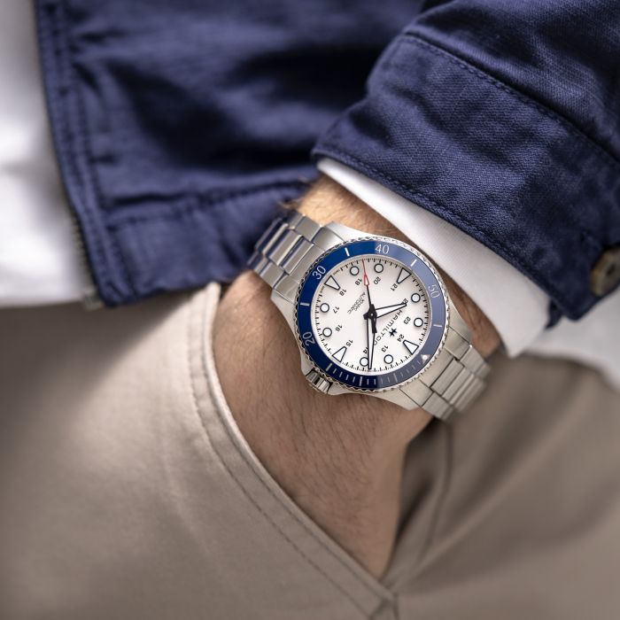 A person wearing a Hamilton Khaki Navy Scuba Automatic Watch with a blue and white face, featuring a blue ceramic bezel and three subdials. The person is dressed in a blue jacket, a white shirt, and beige pants, with their hand casually placed in a pocket, ready for maritime adventures.