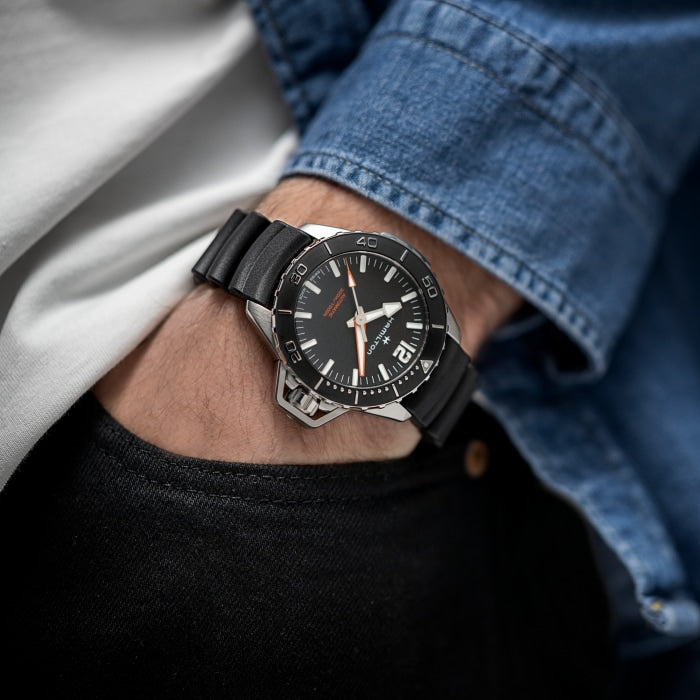 A person wearing a casual outfit of a white shirt and a denim jacket has their hand in the pocket of dark pants. On their wrist is a sleek Khaki Navy Frogman Automatic Watch from Hamilton, featuring a wave-patterned tactical rubber strap, detailed face with white hour markers, and an orange hand—perfect for maritime adventures.