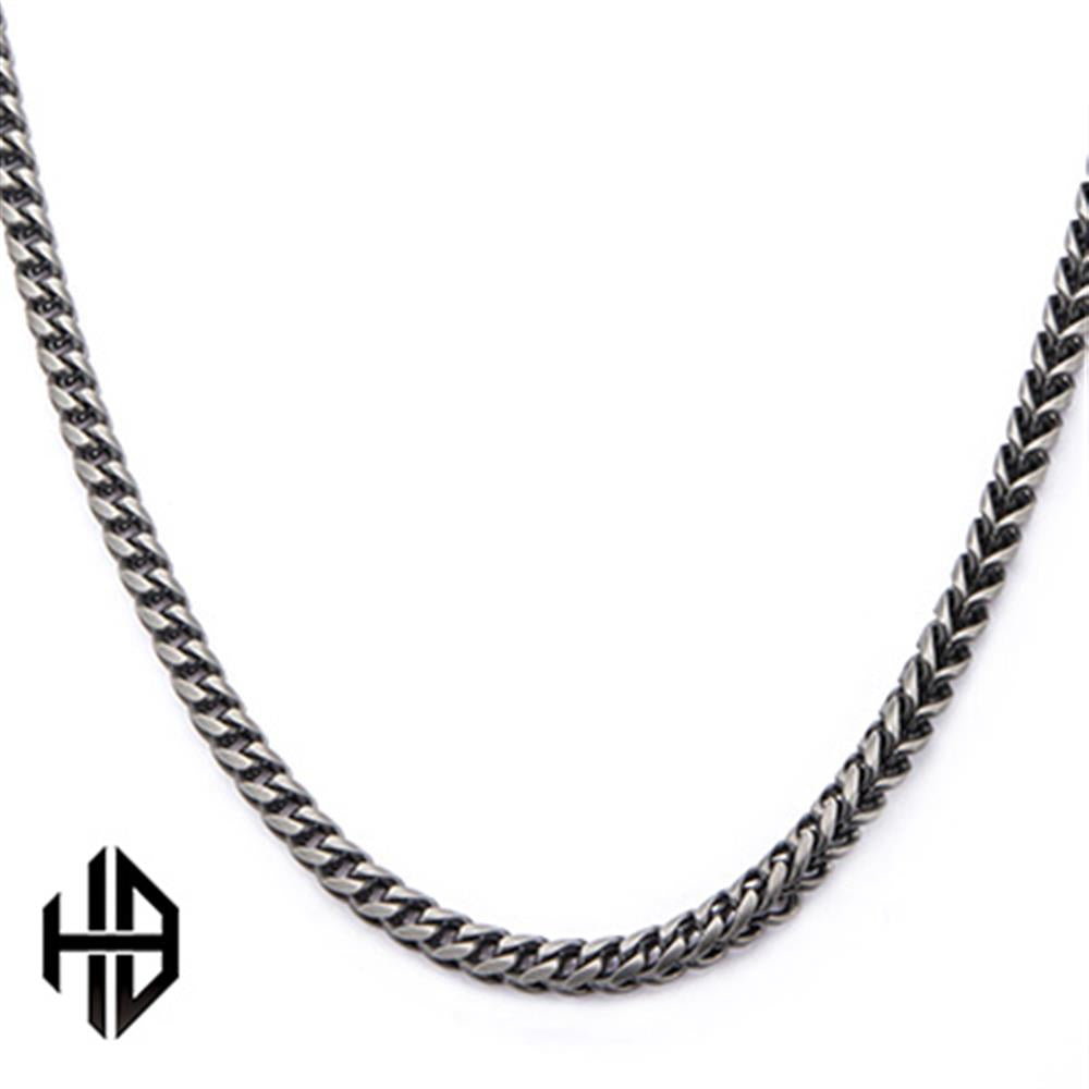 A close-up image of the Hollis Bahringer Men's Stainless Steel Necklace by INOX, showcasing its sleek, silver-toned fox tail link design. The chain forms a continuous loop and is displayed on a plain white background. A small, black geometric logo resembling interconnected letters is visible in the bottom left corner.