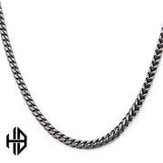 A close-up image of the Hollis Bahringer Men's Stainless Steel Necklace by INOX, showcasing its sleek, silver-toned fox tail link design. The chain forms a continuous loop and is displayed on a plain white background. A small, black geometric logo resembling interconnected letters is visible in the bottom left corner.