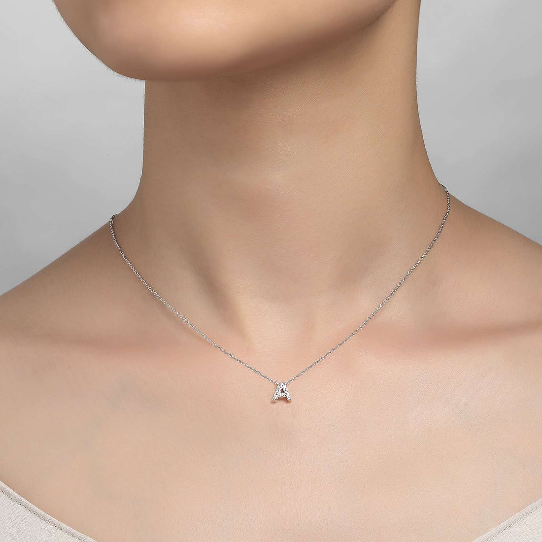 A close-up image of a person wearing the elegant Letter A Pendant Necklace by Lafonn, featuring a delicate silver chain that is adjustable and adorned with a small, triangular pendant encrusted with simulated diamonds. The person's neck and upper chest are visible, devoid of other accessories or clothing details. The background exudes simplicity, rendered in a clean, neutral gray.