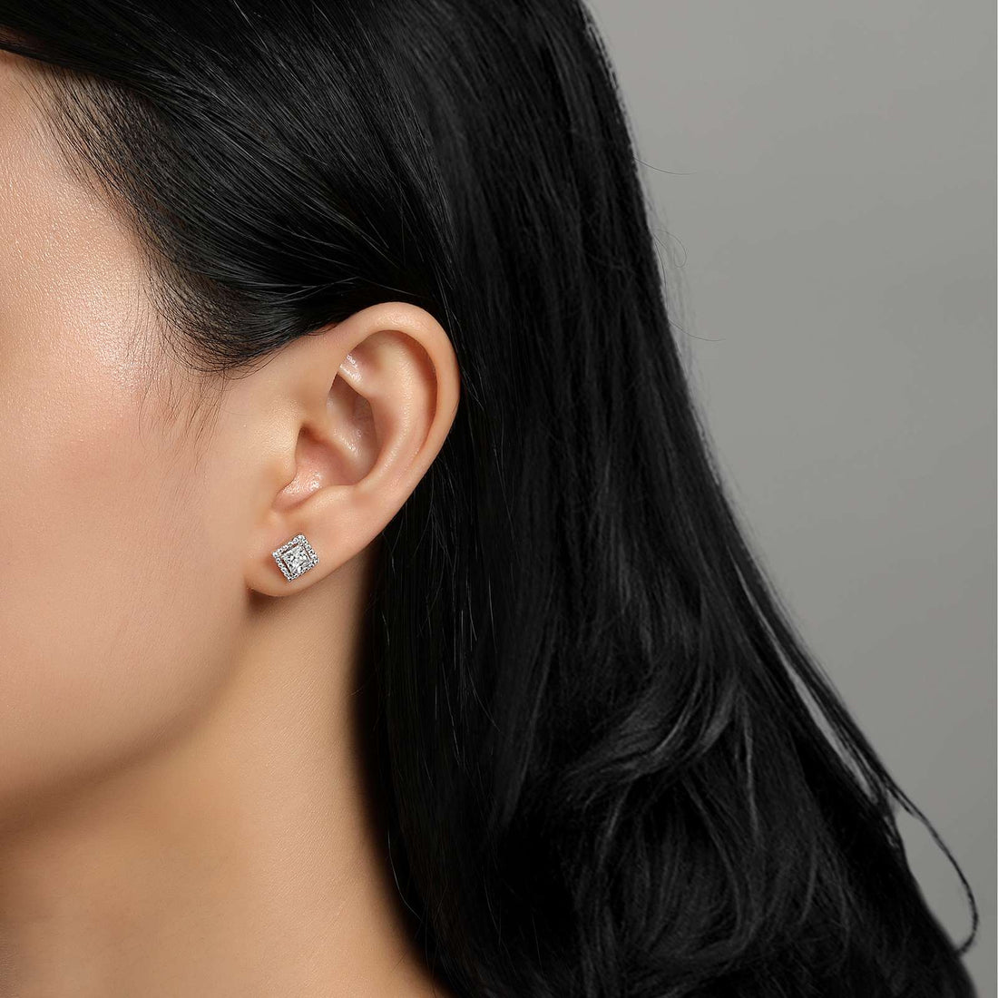 Close-up of a person with long black hair showcasing the 1.50 carats Diamond Stud Earrings by Lafonn in their left ear. The simulated diamond sparkles against their smooth, light skin. The background is plain and gray, emphasizing the stunning jewelry piece from Lafonn.