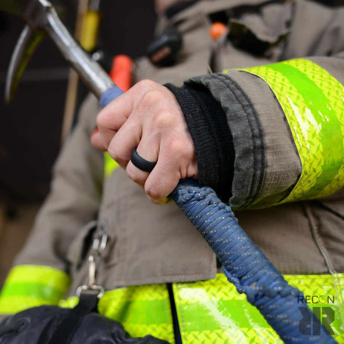 A close-up of a firefighter gripping a rescue tool, wearing protective gear with reflective yellow-green stripes. The handle of the tool is wrapped in blue material. Only part of the firefighter is visible, and he's sporting a Men's Black Silicone Wedding Ring by Baxley Jewelry on his finger. The logo "RECON" appears in the bottom right corner.