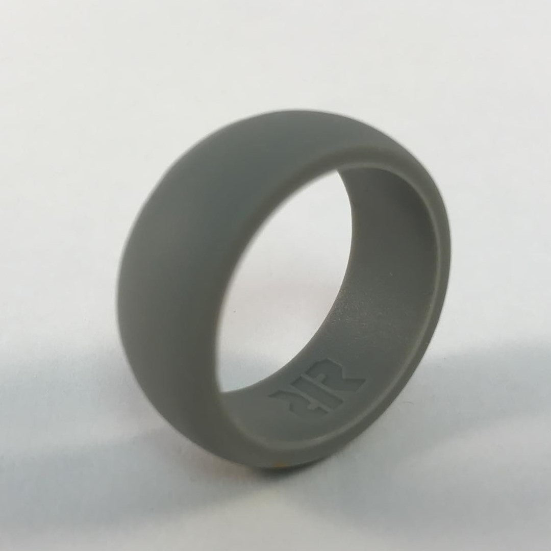 A close-up of a matte gray silicone wedding ring lying on its side on a white surface. Inside the ring, there is a subtle engraving of a logo or symbol consisting of two connected geometric shapes. Crafted from medical grade silicone, this Men's Gray Silicone Wedding Ring by Baxley Jewelry is perfect for active lifestyles.