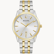 The Bulova Sutton Classic Watch (98B385) by the Bulova Watch Company features a two-tone metal band in silver and gold. It has a round silver watch face with gold hour markers and hands, and includes a date display at the 6 o'clock position. This elegant dress watch is designed with a silver case accented with refined gold details.