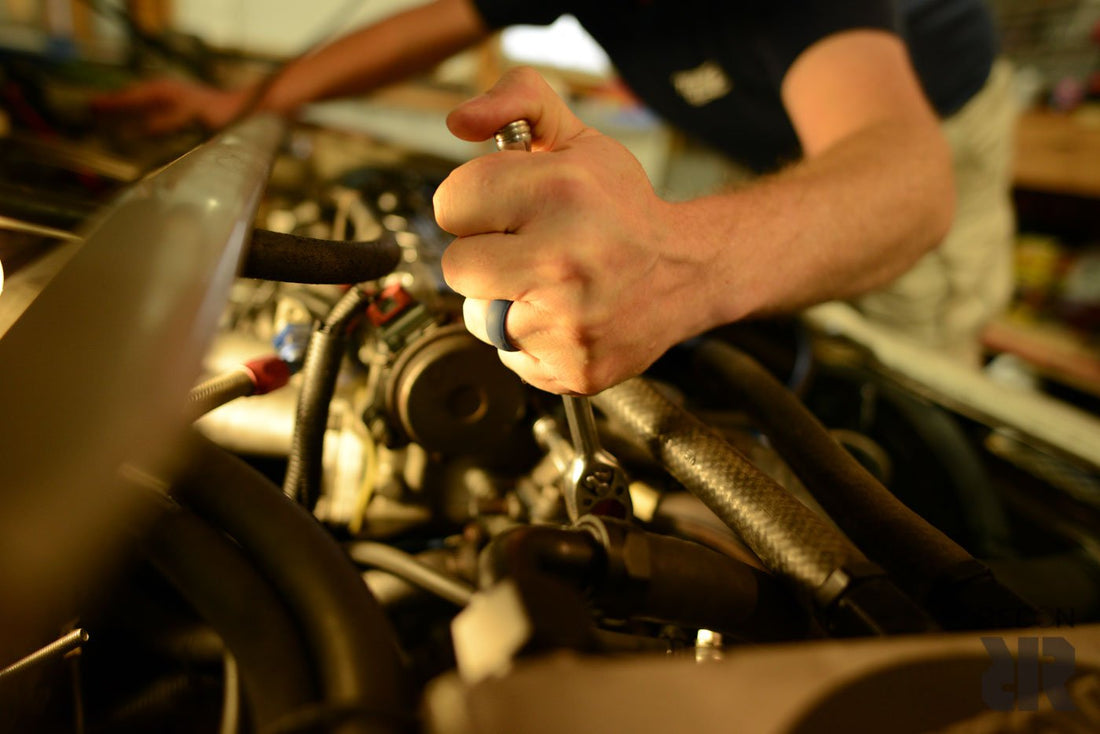 Close-up of a person wearing a Baxley Jewelry Men's Navy Blue Silicone Wedding Ring while using a wrench to turn a bolt in an automotive setting. Only their hand and part of their arm are visible. The background includes various hoses and engine parts, indicating they are working on an engine.