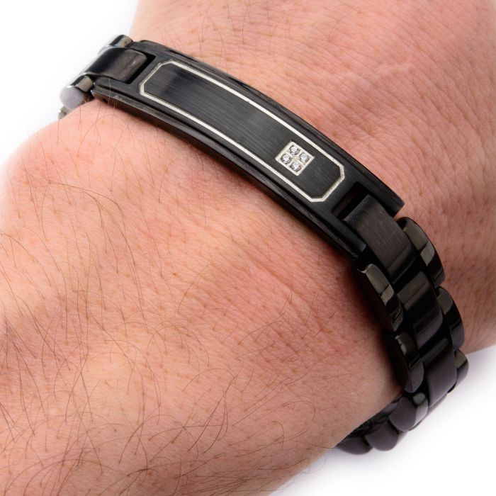 A close-up image captures a wrist adorned with the INOX Men's Stainless Steel Black Plated with Clear CZ ID Plate Link Engravable Bracelet. This sleek, adjustable bracelet (8.25" - 8.75") features a black-plated design and a small, rectangular silver accent in the center. The wearer's hand is visible against a plain white background.