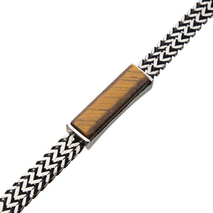 A close-up of the INOX Stainless Steel Double Franco Chain with Tiger Eye Stone ID Link Bracelet, highlighting its intricately braided black and white band. The rectangular Tiger Eye Stone clasp gleams with a shiny, brownish-golden hue and natural streaks, set in a durable metallic frame. The bracelet's robust Double Franco Chain pattern forms an elaborate zigzag design.