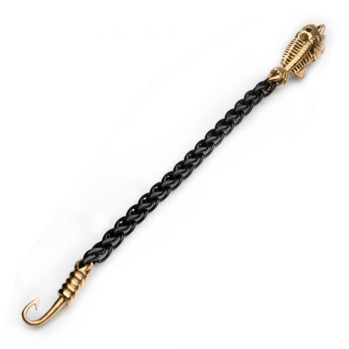 A black braided bullwhip with gold-colored metal accents, featuring a handle intricately designed to resemble a pharaoh's head and ending in a curved hook, is neatly arranged beside the sleek Black Plated Wheat Chain with Gold Plated Fishbone on Hook Clasp Bracelet by INOX against a plain white background.