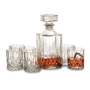 A 7 Piece Crystal Whiskey Set, consisting of six 6-8 oz glasses and a 32 oz decanter partially filled with amber liquid. The decanter comes with a square, faceted stopper, and all pieces feature an intricate cut-glass design. The glasses are arranged around the decanter.