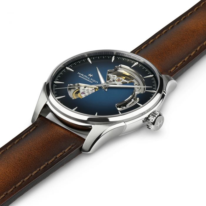 The Hamilton Jazzmaster Open Heart Automatic Watch showcases a smoked blue dial with exposed mechanical movement, metallic indices, and a polished silver case. It features a brown leather band with visible stitching and intricate inner workings displayed through partial cutouts on the face.