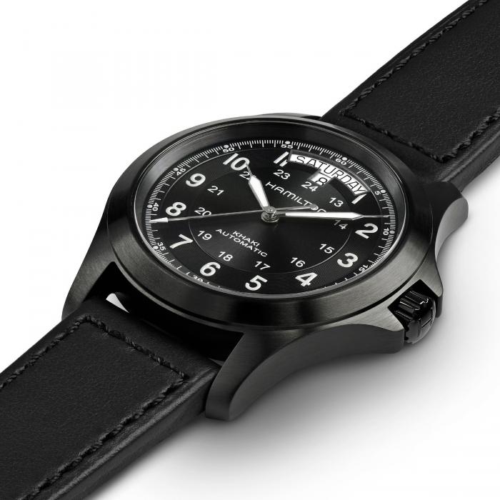 The Khaki Field King Automatic Watch by Hamilton features a black leather strap and a black dial with white numerals, 1-12 and 13-24. The round face has a day and date window at the 12 o'clock position, with the brand name "Hamilton" prominently displayed on the dial to highlight its military heritage as one of the finest field watches.