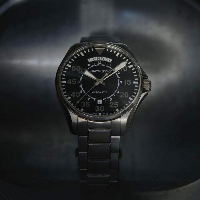 A close-up of the Khaki Aviation Pilot Day Date Automatic Watch by Hamilton. The pilot watch features a metallic band and a dark dial with large, luminous numerals and hour markers. The day and date are displayed at the 12 o'clock position, with the word "Hamilton" prominently visible below the day display.
