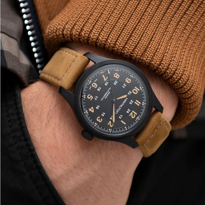 A person wearing a brown sweater is showcasing a Hamilton Khaki Field Titanium Automatic Watch, featuring a tan leather strap and a black watch face. This 42mm wristwatch displays both 12-hour and 24-hour formats with notable brand detailing. The person's hand is tucked into a black pocket.
