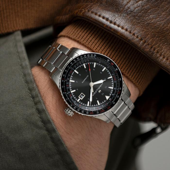 Close-up of a person wearing a Hamilton Khaki Aviation Converter Automatic Watch in stainless steel with automatic movement and a black dial. The watch features a date window at 3 o’clock, a rotating bezel with various markings, and a metal bracelet. The person is dressed in a brown knitted sweater and leather jacket.