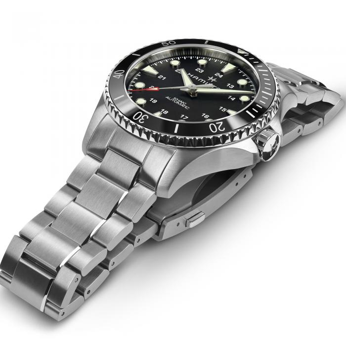 A sleek, silver stainless steel wristwatch with a black ceramic bezel and dial, featuring luminescent hour markers and hands. The 43mm case houses the Khaki Navy Scuba Automatic movement by Hamilton and is partially folded, showcasing its bracelet-style metal band and crown detailing.