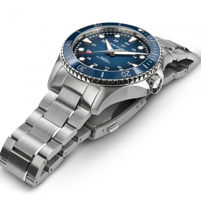 Displayed is the Hamilton Khaki Navy Scuba Automatic Watch, a luxury timepiece featuring a 43mm stainless steel case and water resistance of 300 meters. The watch showcases a blue dial and bezel, paired with a bracelet-style band. It includes silver-tone indices, hands with a date window, minute markers on the bezel, white hour markers, and small red accents for added detail.