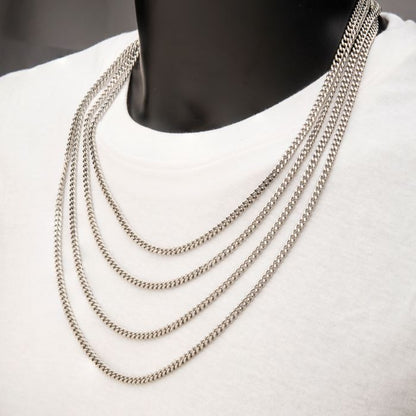 A close-up image of a person wearing a white shirt and multiple layered silver chains around their neck. The chains, including the 4mm Steel Diamond Cut Curb Chain Necklace by INOX, vary slightly in length and have a uniform, braided design. The background is out of focus.