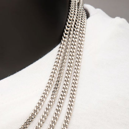 Close-up shot of a person wearing several INOX 4mm Steel Diamond Cut Curb Chain Necklaces, each measuring 22", over a white shirt. The chains have a similar diamond cut curb link design but vary in length, creating a layered effect against the gray background.