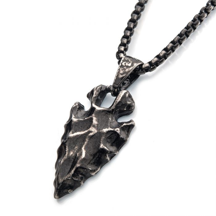A close-up image of an INOX Stainless Steel Gun Metal Plated Chiseled Arrowhead Pendant | 24" featuring a rugged, arrowhead-shaped pendant made of dark, textured metal. The pendant hangs from a thick, black box chain with linked segments, and the bail connecting the pendant to the chain has an ornamental design.